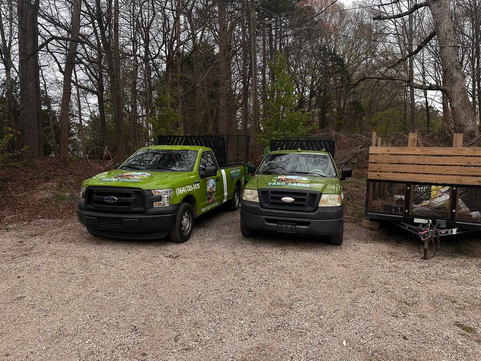 Two green pickup trucks parked on a gravel lot beside a wooden trailer, with leafless trees behind.