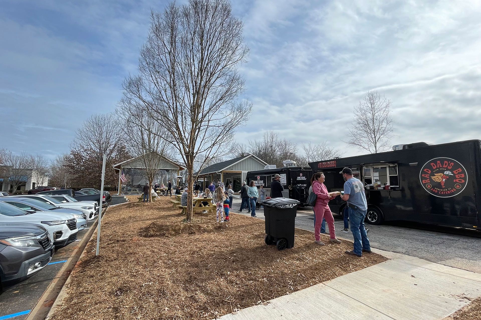 People standing outside a storefront beside parked cars and leafless trees on a cloudy day