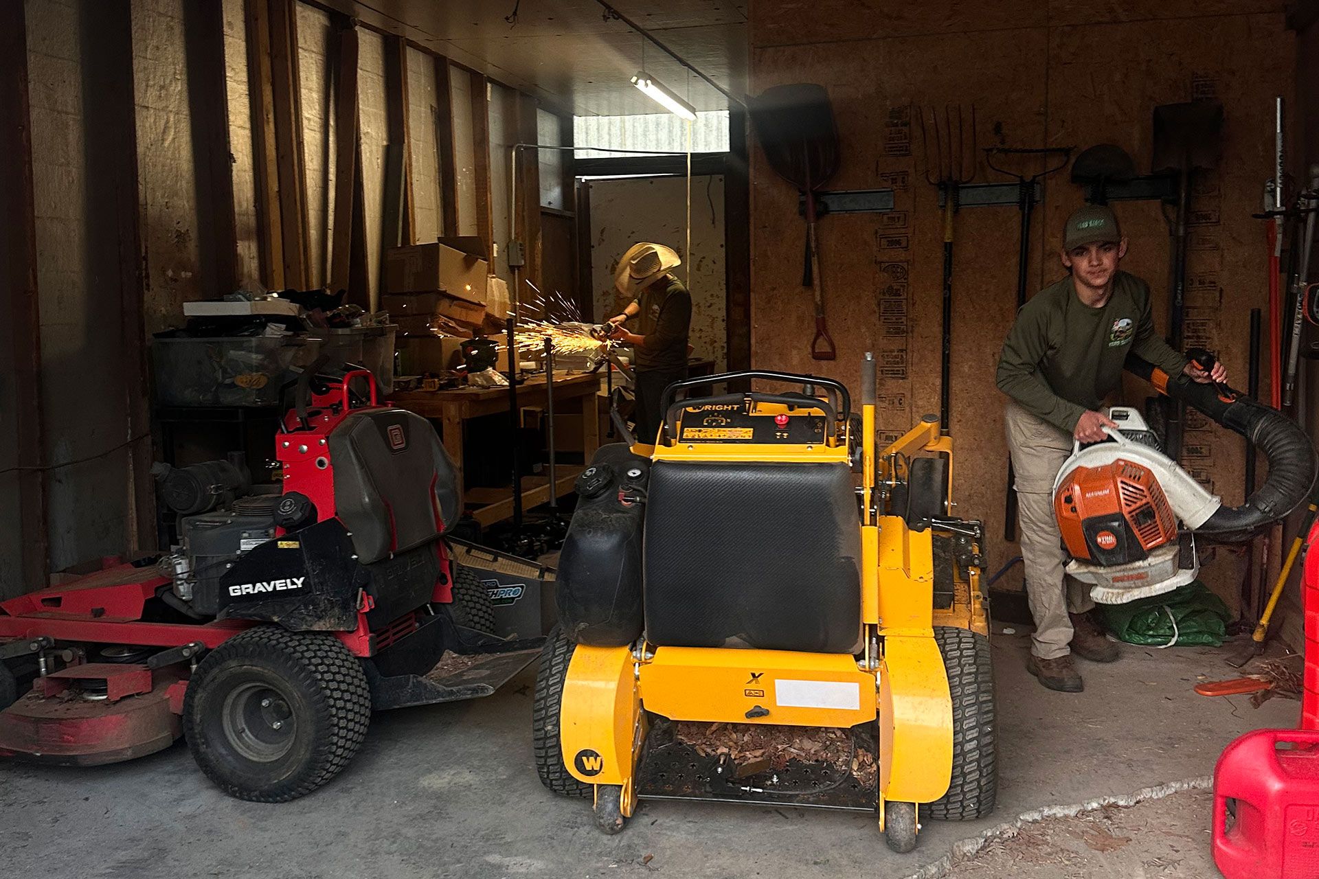 Two riding mowers and a yellow skid steer in a dim garage; a person stands beside an orange chainsaw.