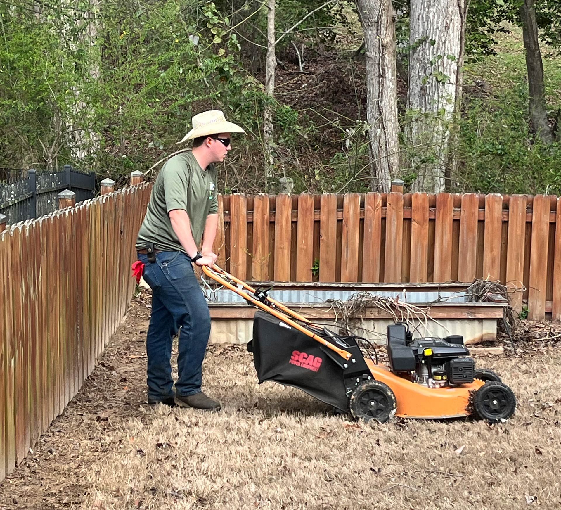 Person mowing dry grass with a push lawn mower beside a wooden fence in a backyard