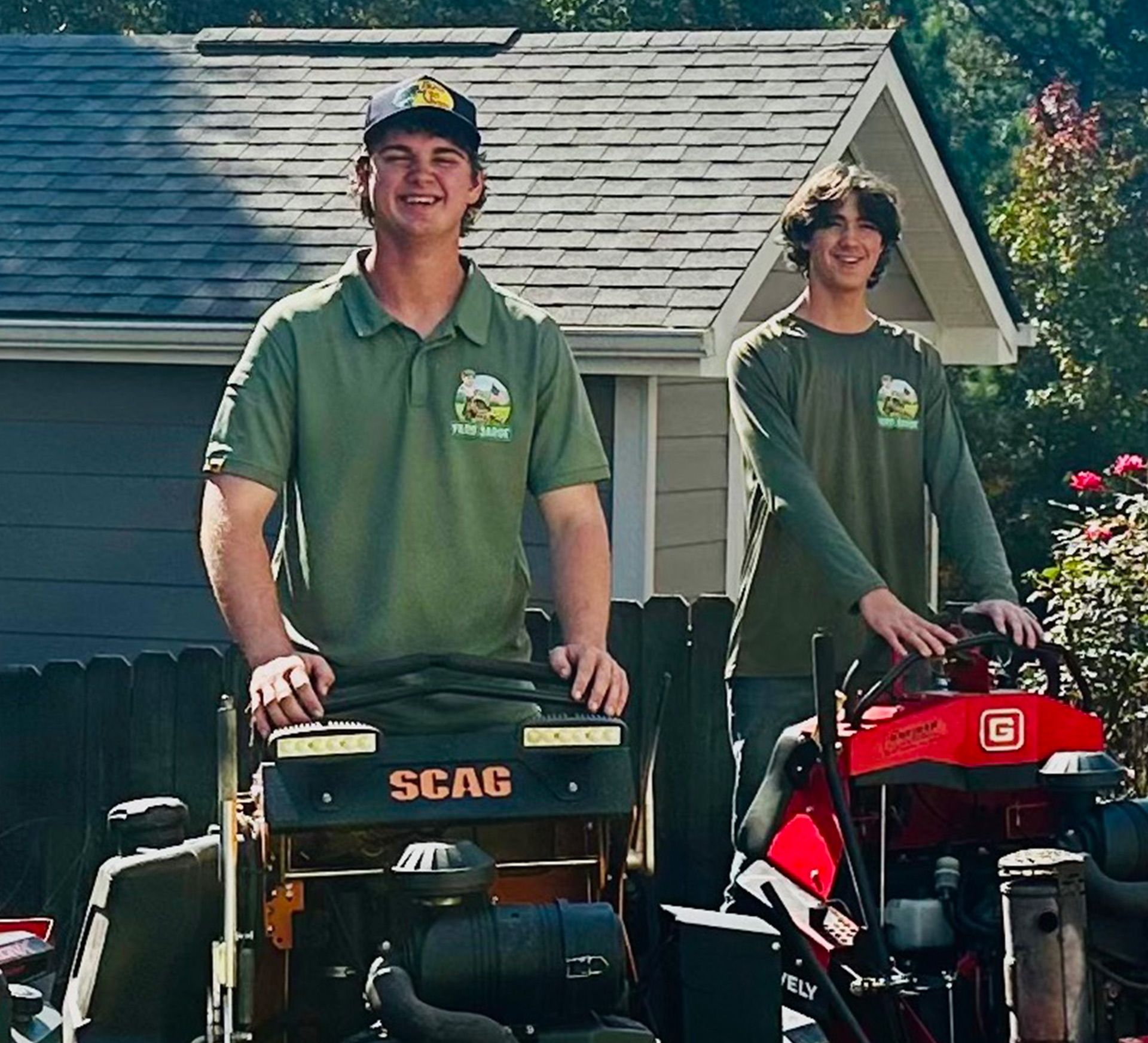 Two workers in green uniforms stand on lawn mowers in front of a house