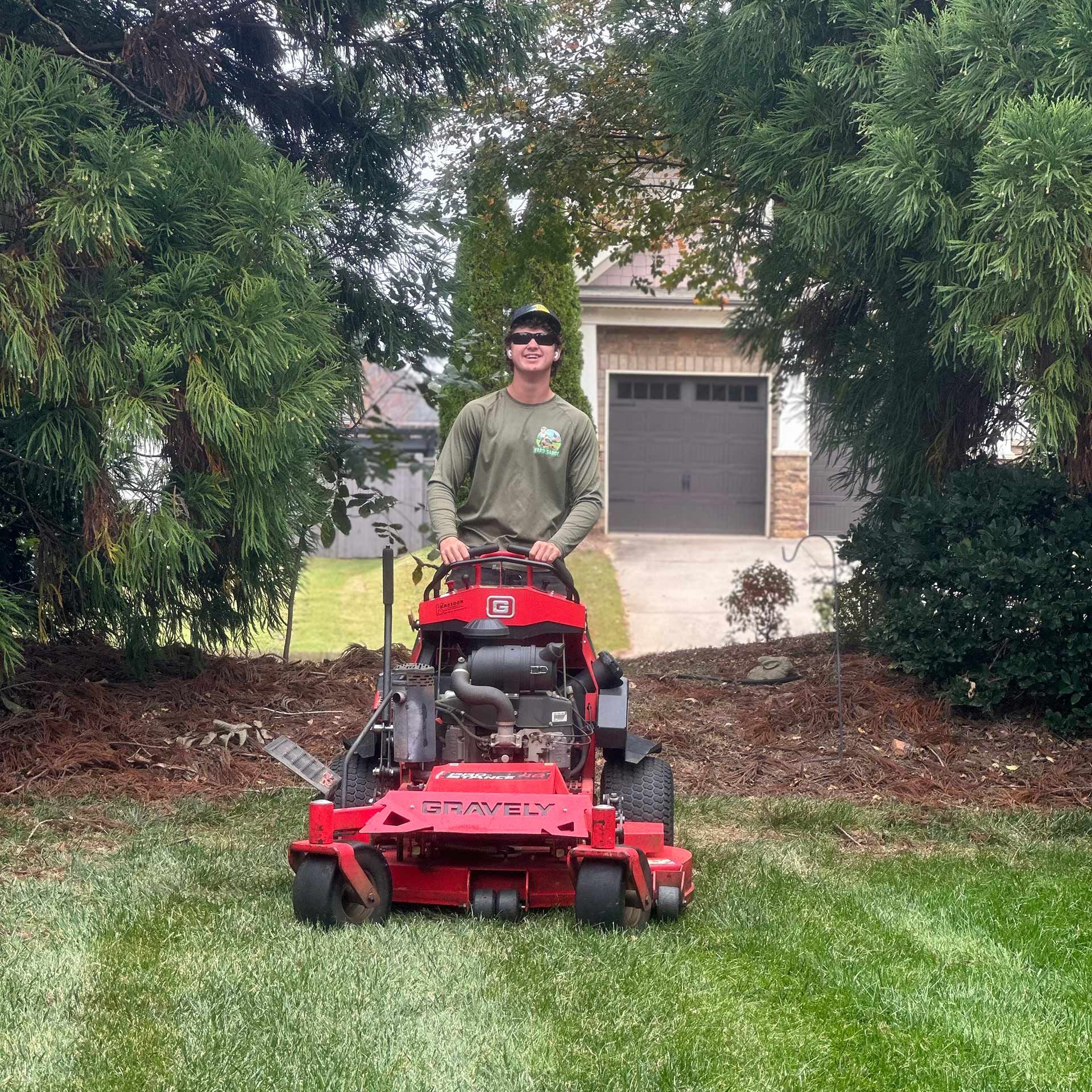 Man standing behind a red riding mower on a lawn between trees and a house