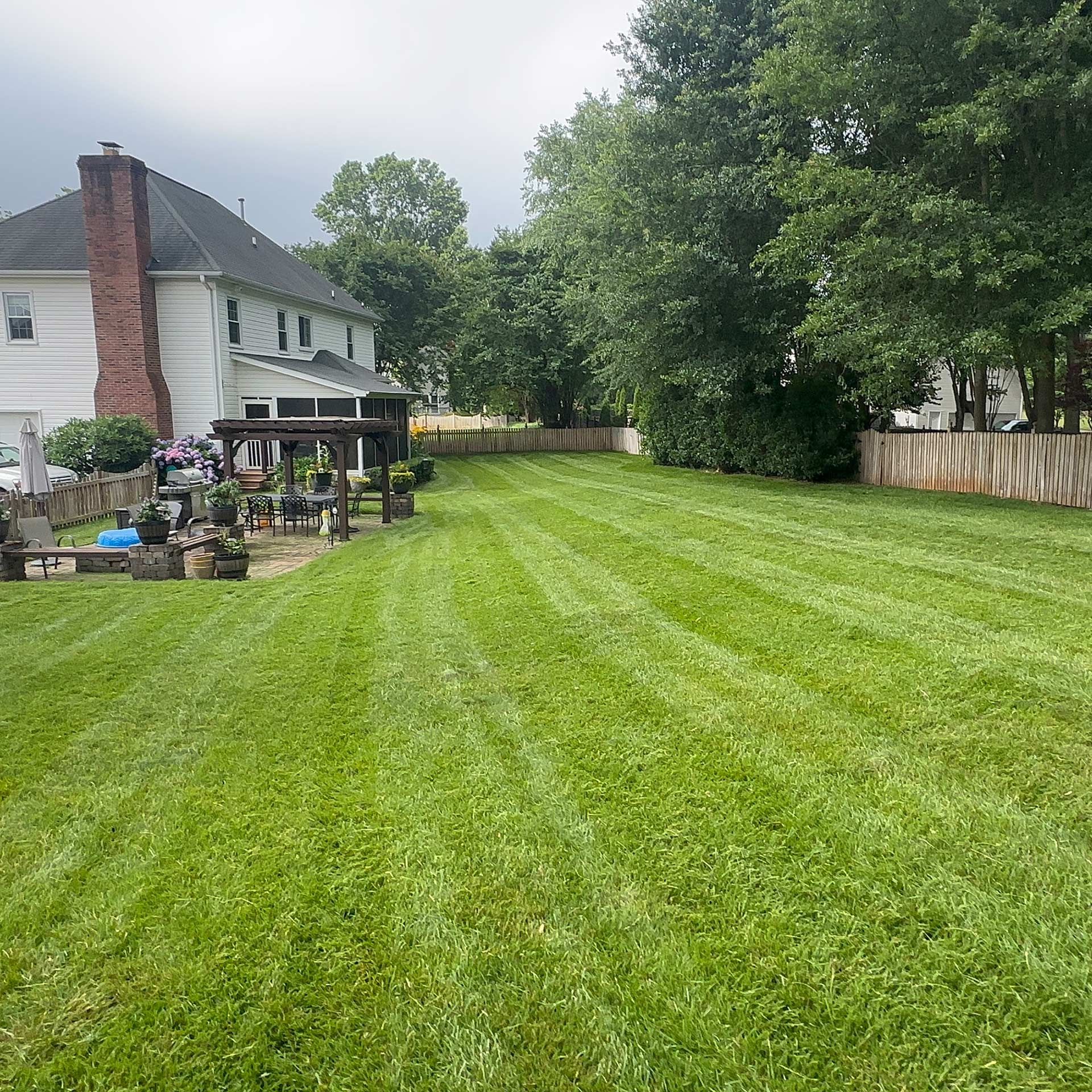 Backyard lawn beside a white house, with patio furniture, brick chimney, and a wooden fence lined with trees