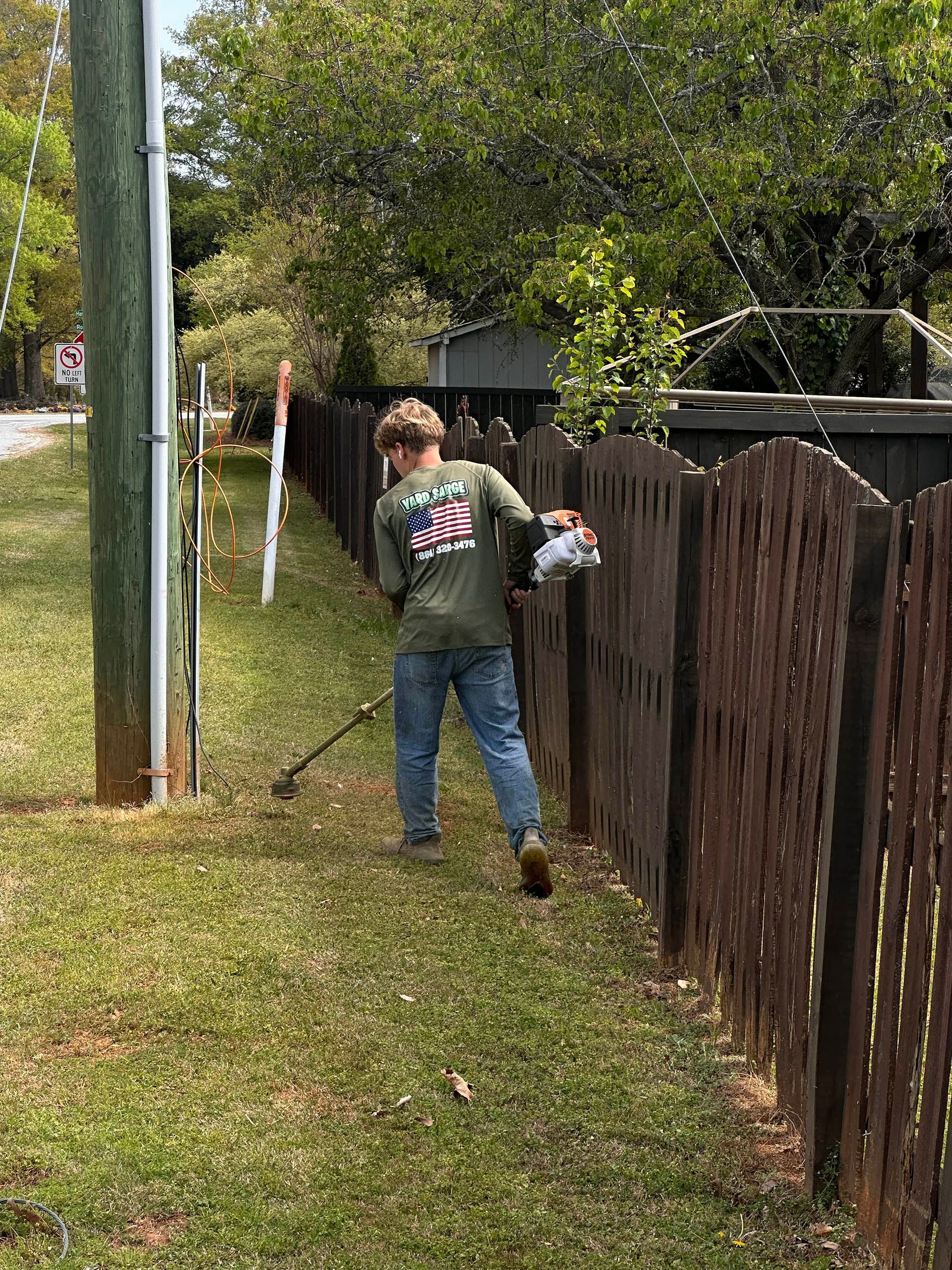 Person walking along a grassy path beside a wooden fence, carrying an item near a utility pole.