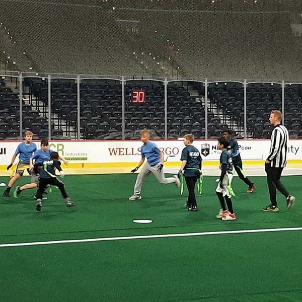 Kids playing indoor football on green turf near the goal, with a referee watching.