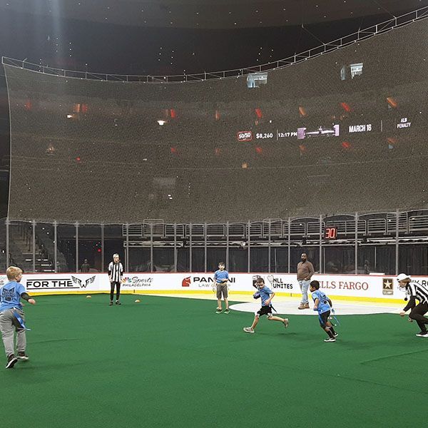 Indoor football game on green turf, with players in blue and white running near the boards under stadium lights.