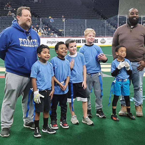 Five boys and two adults posing on an indoor sports field, wearing matching blue shirts.