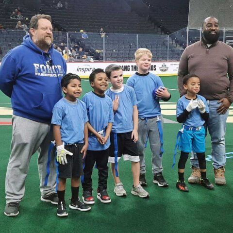 Group of children in blue shirts posing with two adults on an indoor sports field, smiling during a team event.