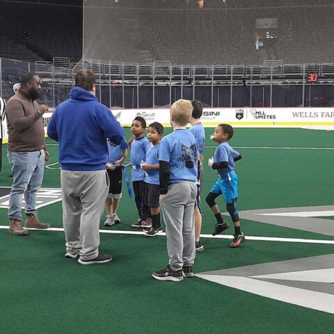 Children in blue shirts gather with coaches on an indoor turf field, likely before a game.
