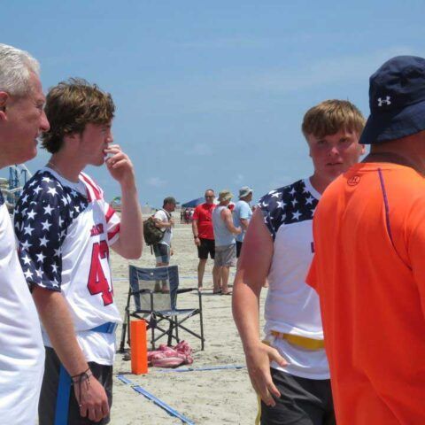 People on a beach in patriotic jerseys, standing and talking near folding chairs under a clear blue sky