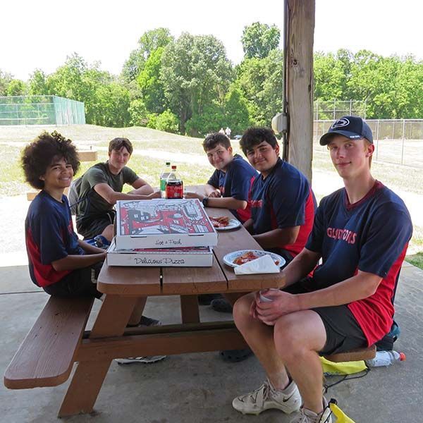 Five people in navy shirts sit around a picnic table outdoors with pizza boxes and soda bottles.