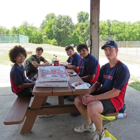 Five teammates in red and navy jerseys sit at a picnic table outdoors, with a pizza box in front of them.