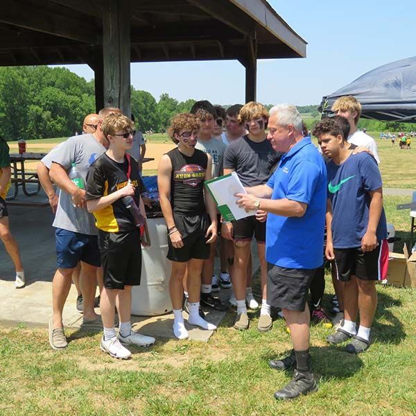 Group of people gathered outdoors around a man in a blue shirt holding papers under a pavilion.