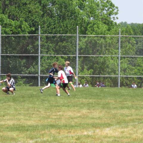 Players chasing a football on a grassy field near a chain-link fence and trees