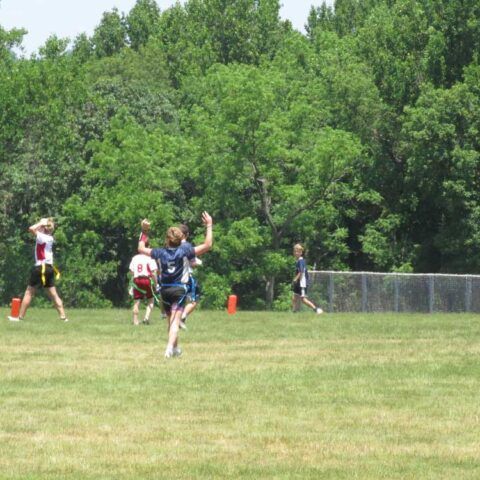 People playing football on a grassy field near a tree line and chain-link fence