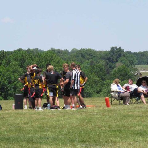 Youth sports team huddles on a grassy field near seated spectators and a wooded backdrop.