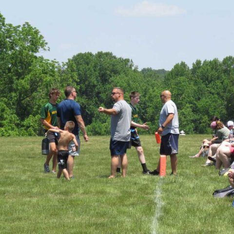 People standing and sitting on a grassy field near a white line, with trees in the background.