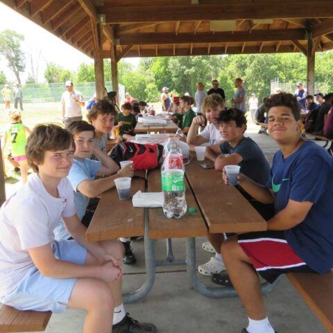 Group of people sitting at picnic tables under a pavilion, sharing drinks and snacks at an outdoor gathering