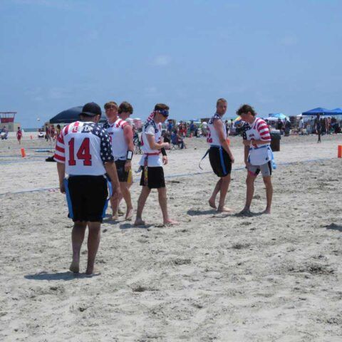 Beach football players in red-and-white jerseys gather on a sandy field under a clear blue sky.