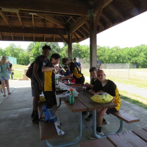 People gathered at a park pavilion picnic table, eating and chatting with a grassy field in the background