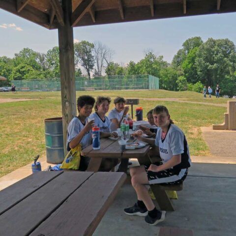 Four people sit at a picnic table under a pavilion, eating and drinking on a sunny grassy field.