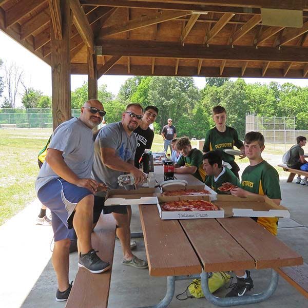 People gathered at a picnic shelter around pizzas and drinks, posing at a wooden table.