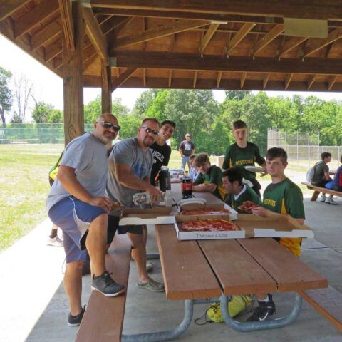 People sitting at a picnic table under a pavilion, sharing pizza and chatting outdoors.