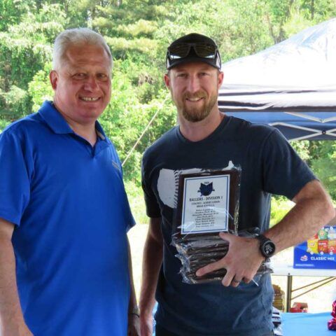Two men smiling outdoors; one holds a plaque in front of a tent and greenery.