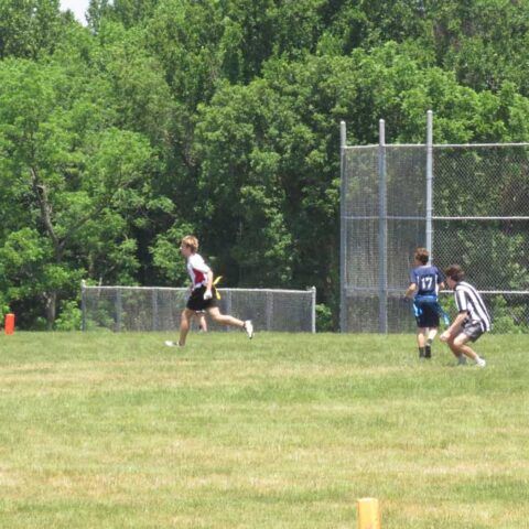 Three people running on a grassy field near a chain-link backstop, with trees in the background.