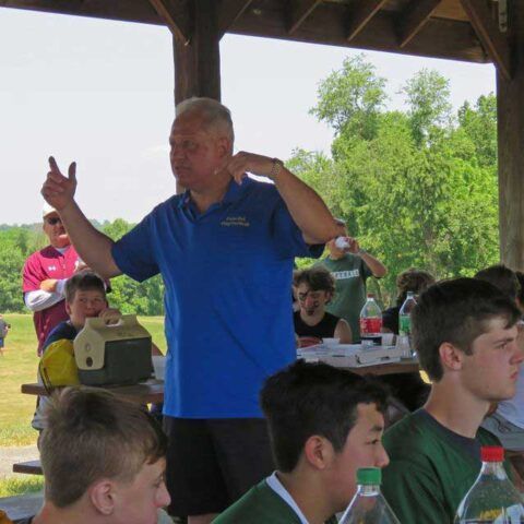 Man in blue polo speaking under a pavilion at a picnic with people seated at tables