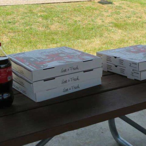 Pizza boxes stacked on a picnic table beside a soda bottle, with grass in the background