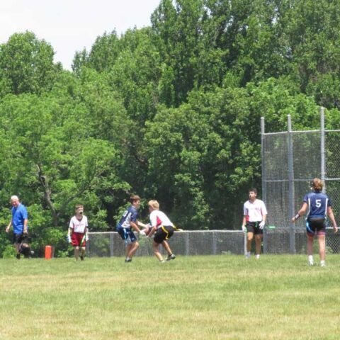 Players in white and blue jerseys compete on a grassy field near trees and a fence.