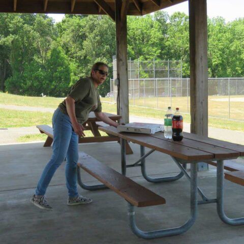 Person arranging a picnic table under a pavilion beside a baseball field, with soda bottles on the table