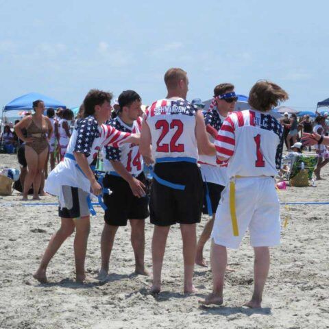 People in patriotic shirts standing on a beach in a circle, holding each other and talking.