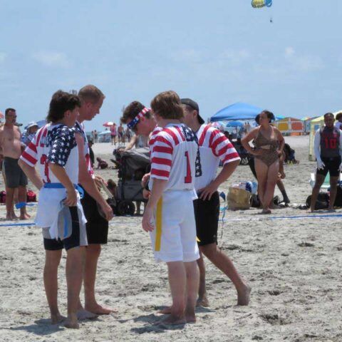 Four people in red, white, and blue striped shirts stand on a sandy beach in a circle.