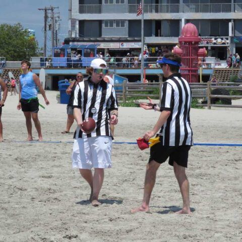 Two referees in black-and-white stripes stand on a sandy beach, one holding a ball and the other a flag.