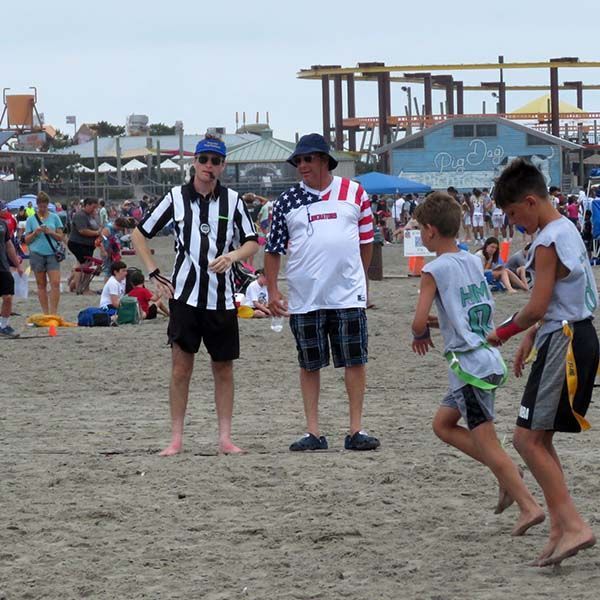 Children and two adults stand on a sandy beach, watching football at a seaside event.