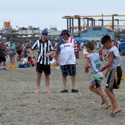 People in sports jerseys playing football on a sandy beach with spectators and rides in the background