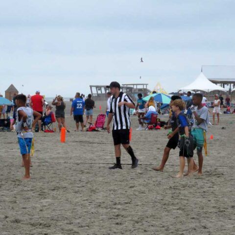 People playing football on a sandy beach, with a referee in black-and-white stripes nearby.