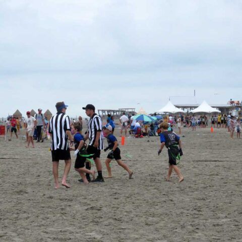People in black-and-white striped shirts play on a sandy beach at an outdoor event with tents in the distance.