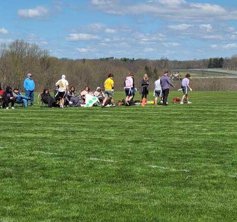 People gathered on a grassy field outdoors under a partly cloudy sky, with cones and a line of participants.