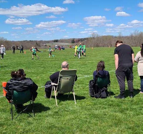 People watching a football game on a sunny grassy field under a blue sky