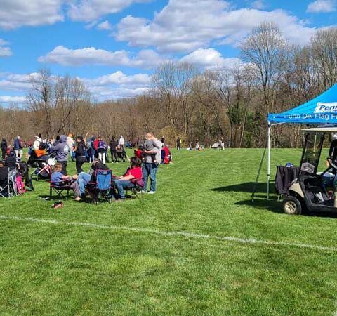 People seated on a grassy field at an outdoor gathering, with a blue tent and golf cart nearby under a cloudy sky