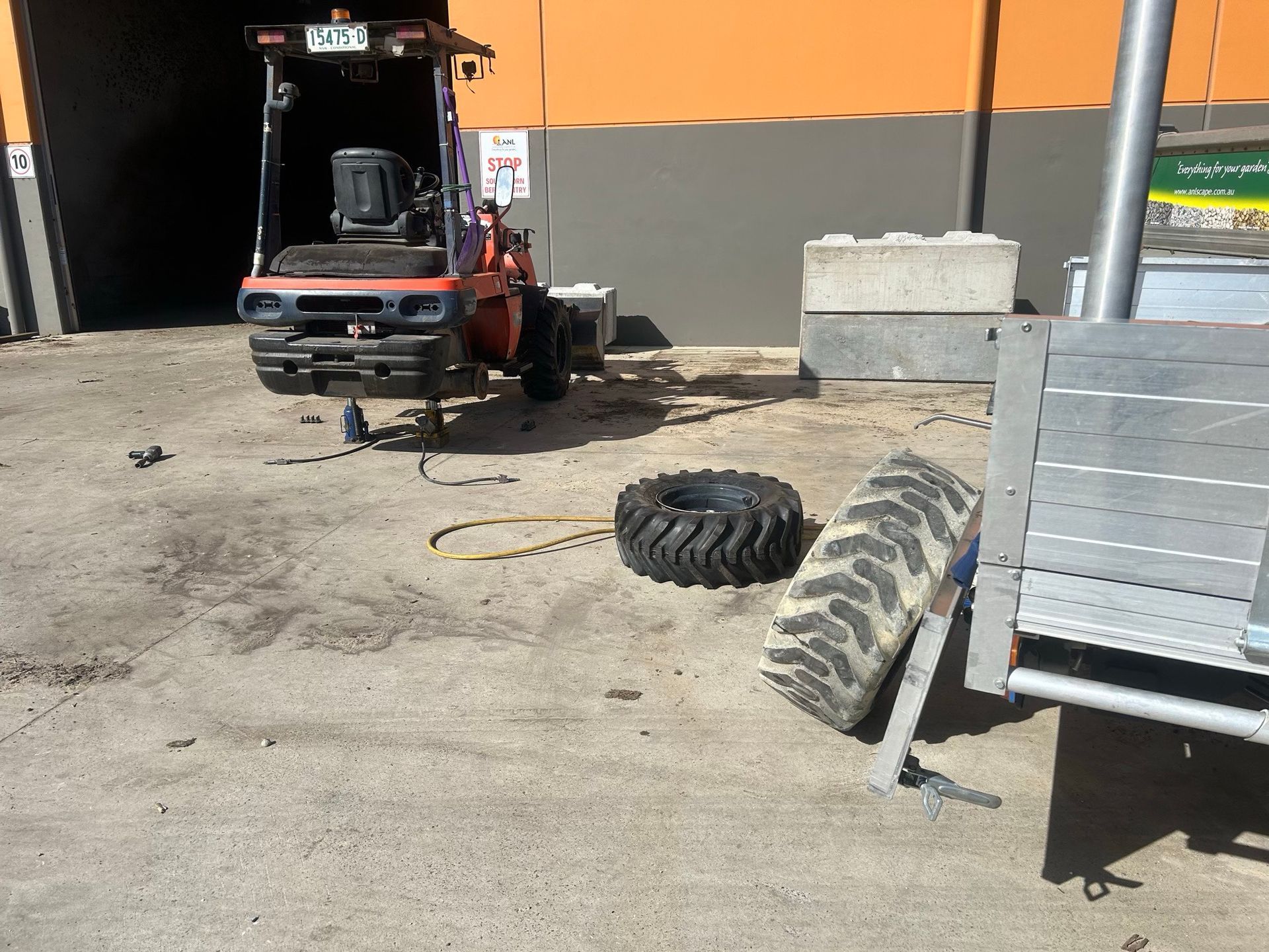 Small red forklift in a yard with two loose tires near a gray trailer and orange building wall.