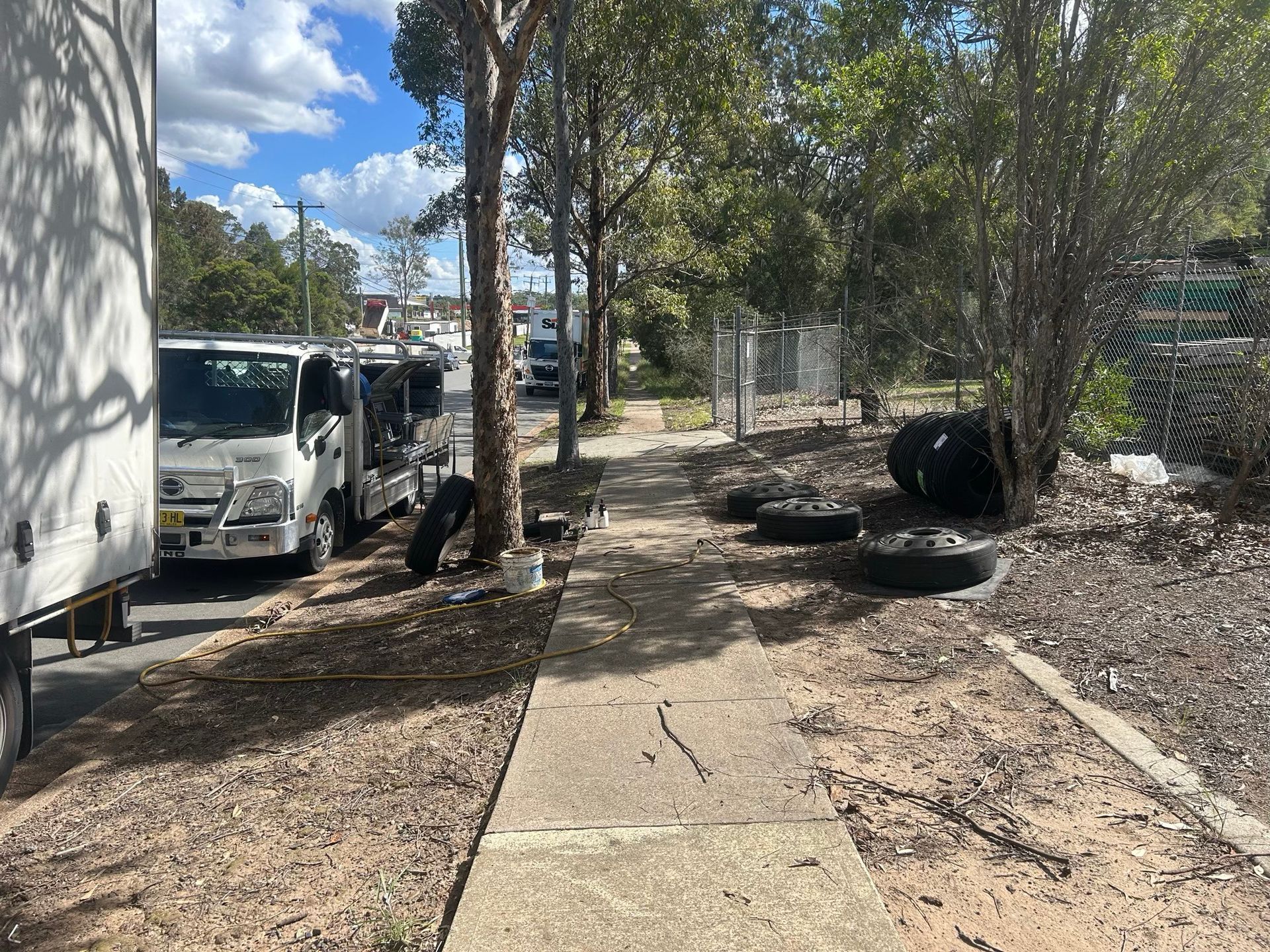 Sidewalk beside parked white vans and trees, with tire stacks along a dirt path under a sunny sky.