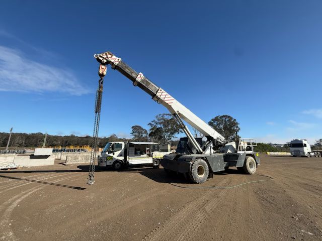 Crane truck lifting a long pole next to a white pickup on a dirt lot under a clear blue sky