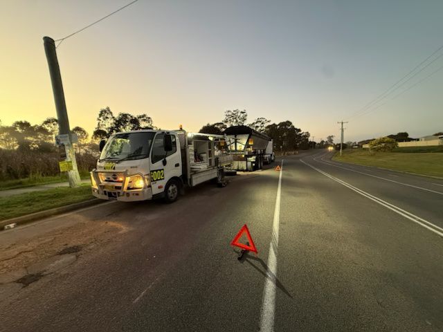 Tow truck with hazard triangle stopped on a road at dusk, towing a truck ahead