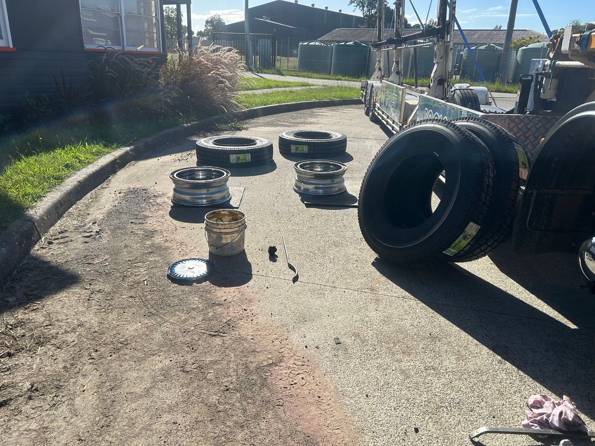 Tires and rims laid out on a sunlit driveway beside a trailer and parked tires.