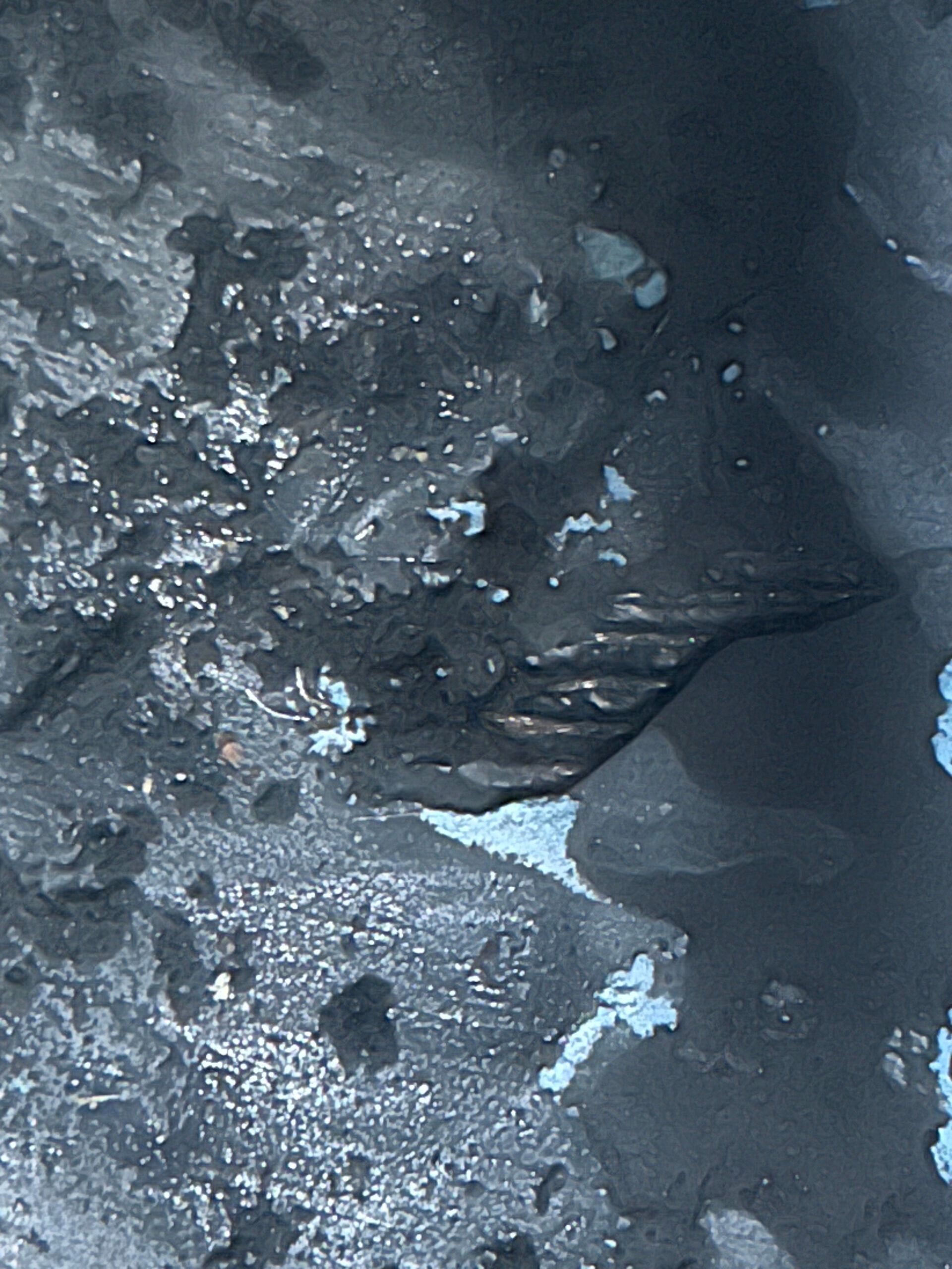 Dark, icy close-up of rough gray rock or frozen surface with patches of white snow and blue ice