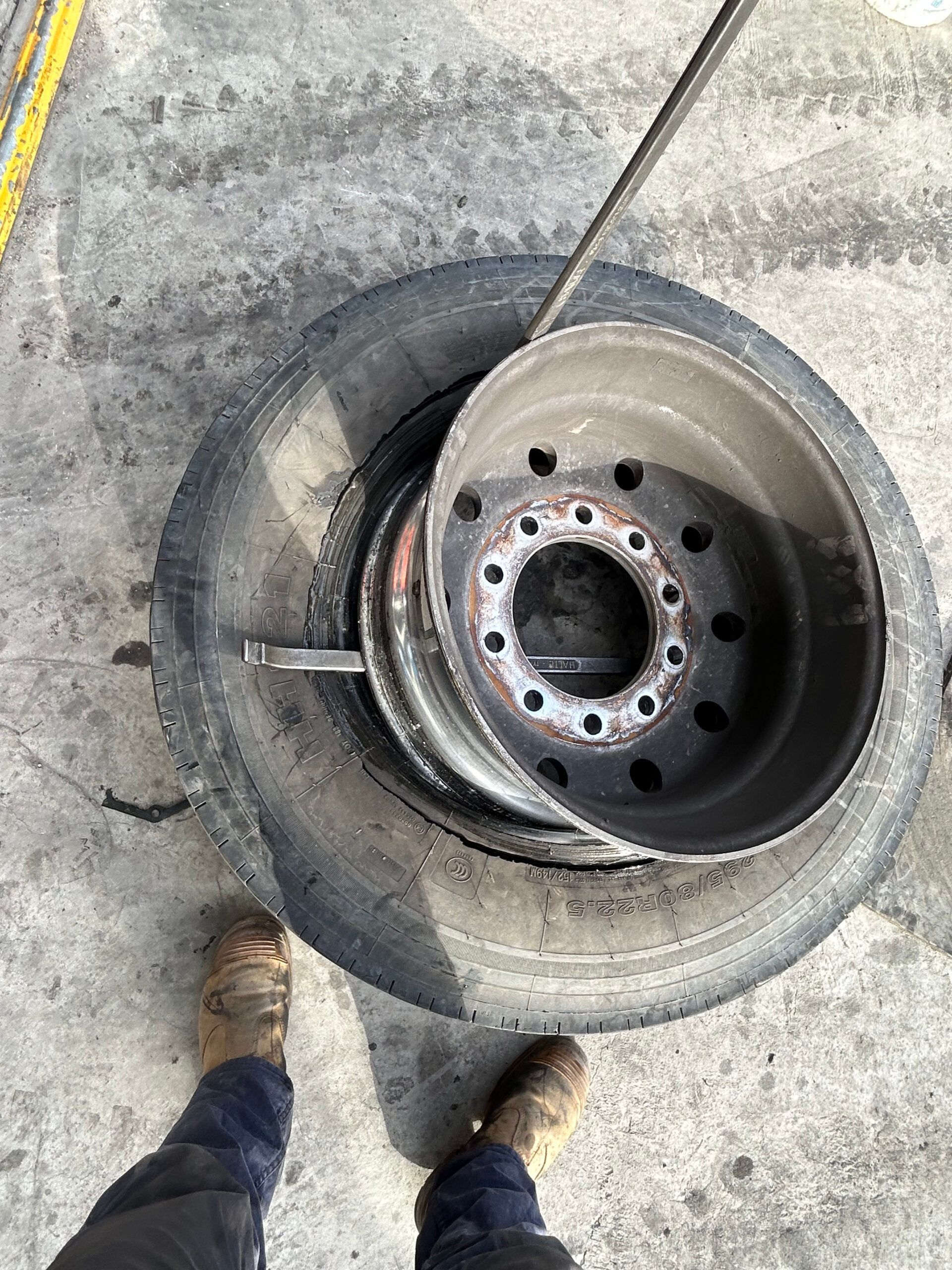 Car tire and wheel rim lying on concrete, with a person’s shoes visible at the bottom.